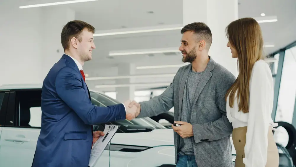 A broker shaking hands with a client beside a new car